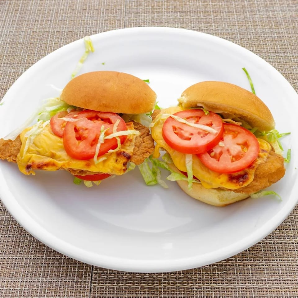 Two chicken sandwiches with melted cheese, lettuce, and sliced tomatoes on a white plate.