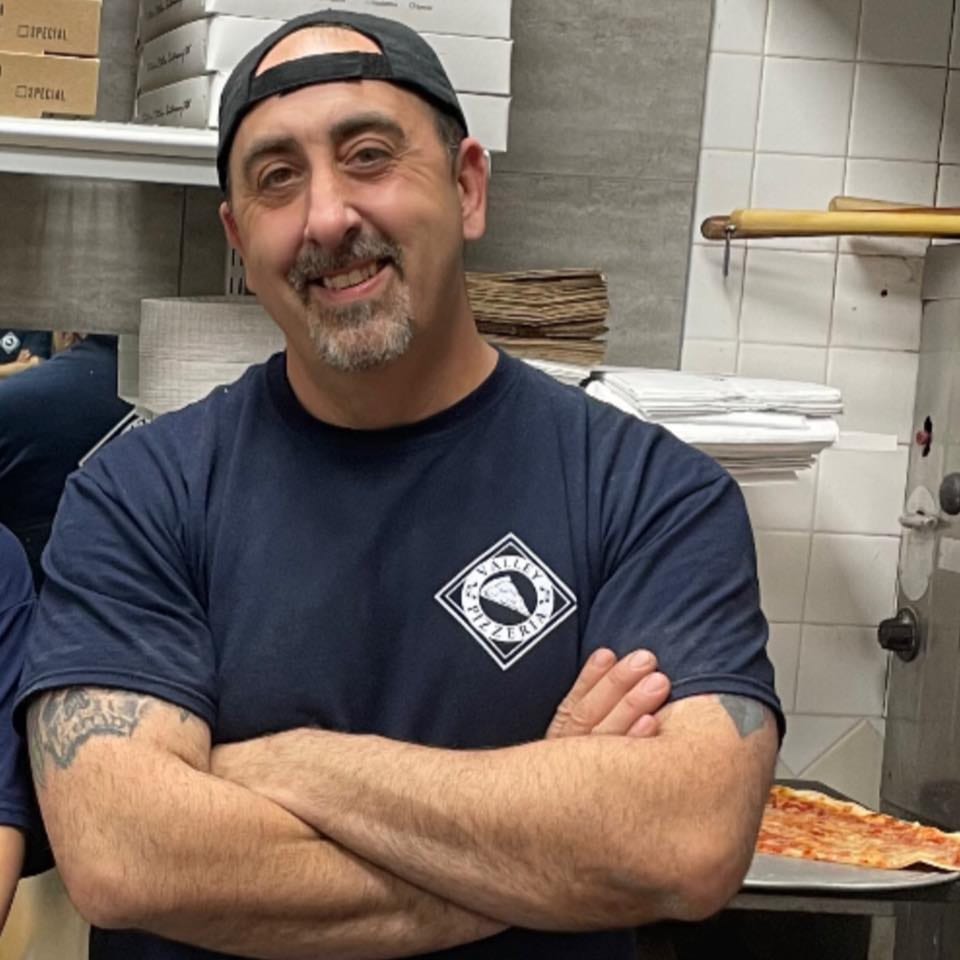 Smiling Napoli's Pizza chef with crossed arms in a kitchen, a pizza crust visible.
