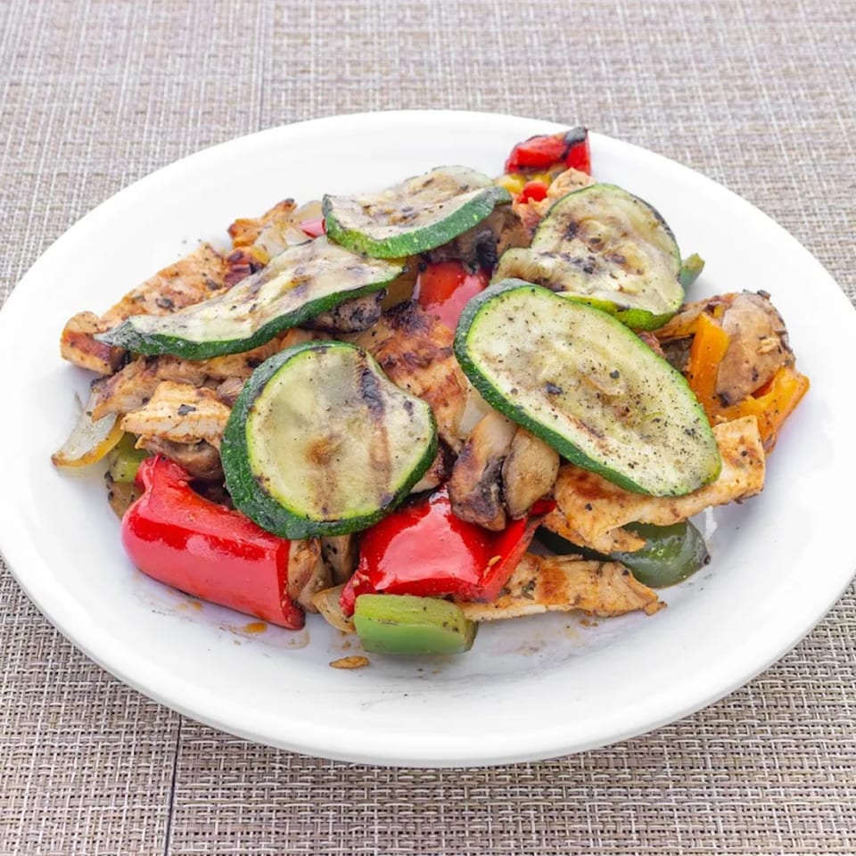Grilled chicken and vegetables, including zucchini, red and green bell peppers, and mushrooms, on a white plate.