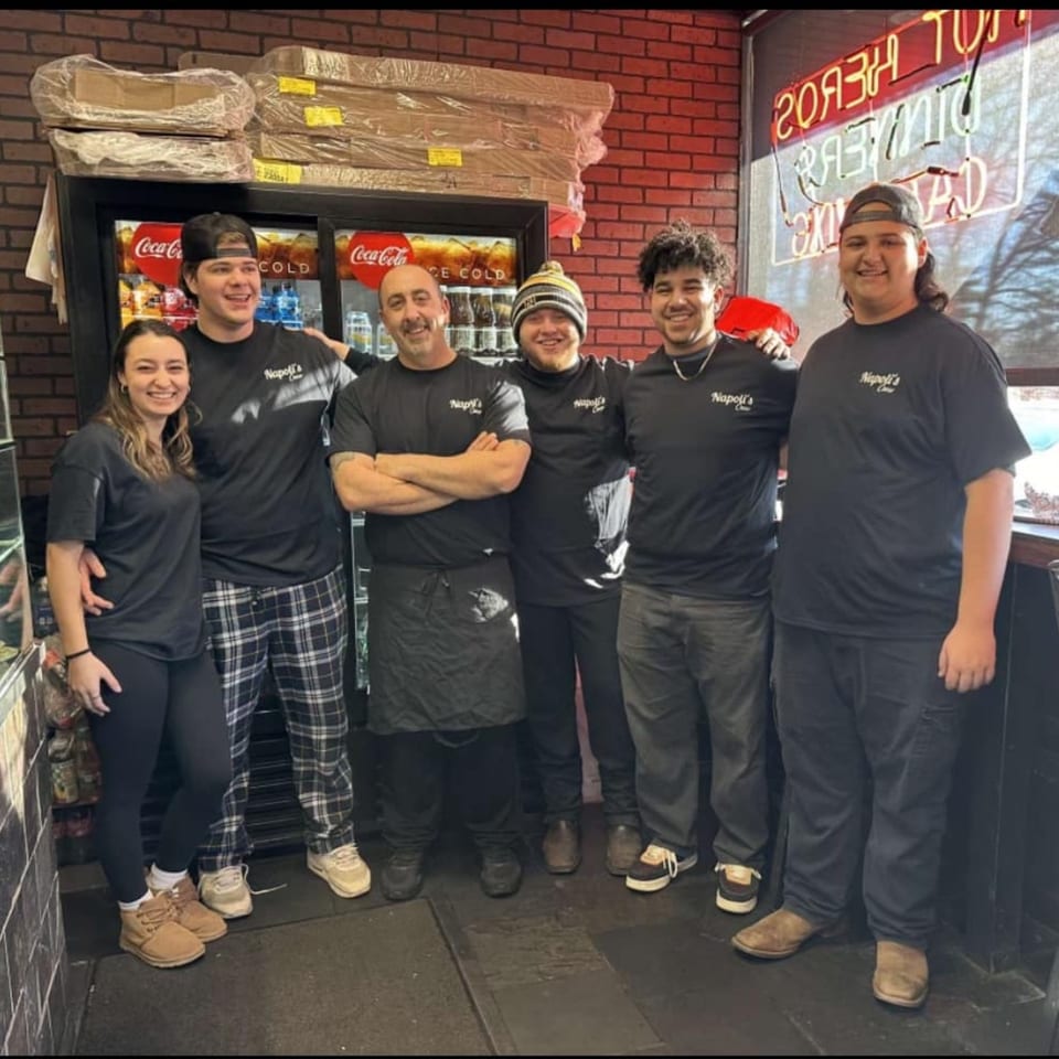 Six smiling Napoli's Pizza & Catering staff members in black shirts stand together in the restaurant.